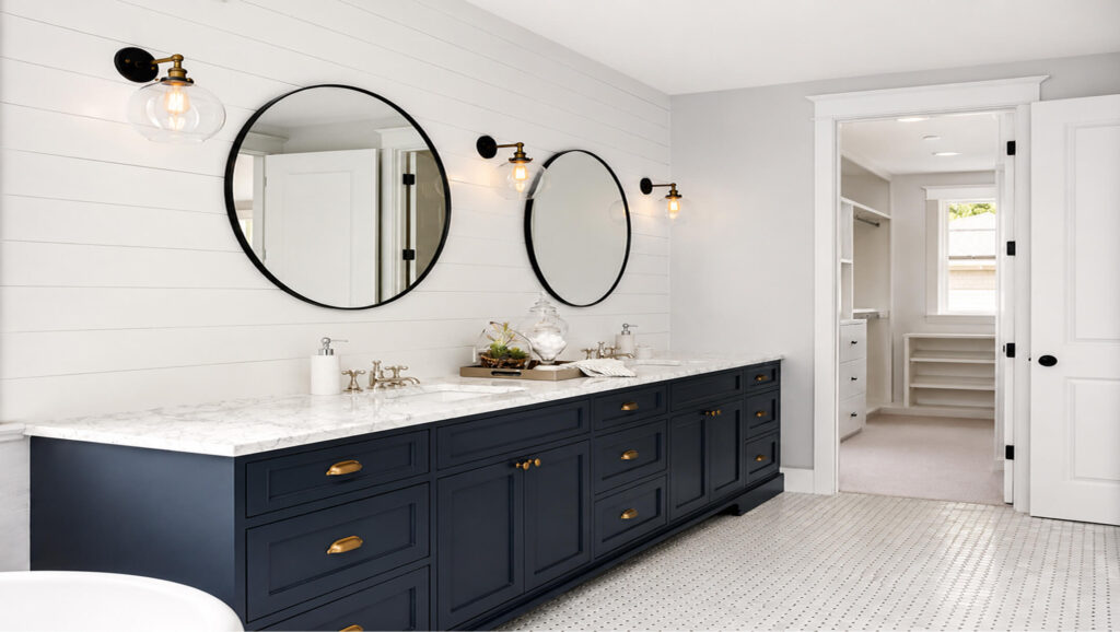 Bathroom with blue cabinets and gold handles. The counters are marble, and the mirrors are oval-shaped.
